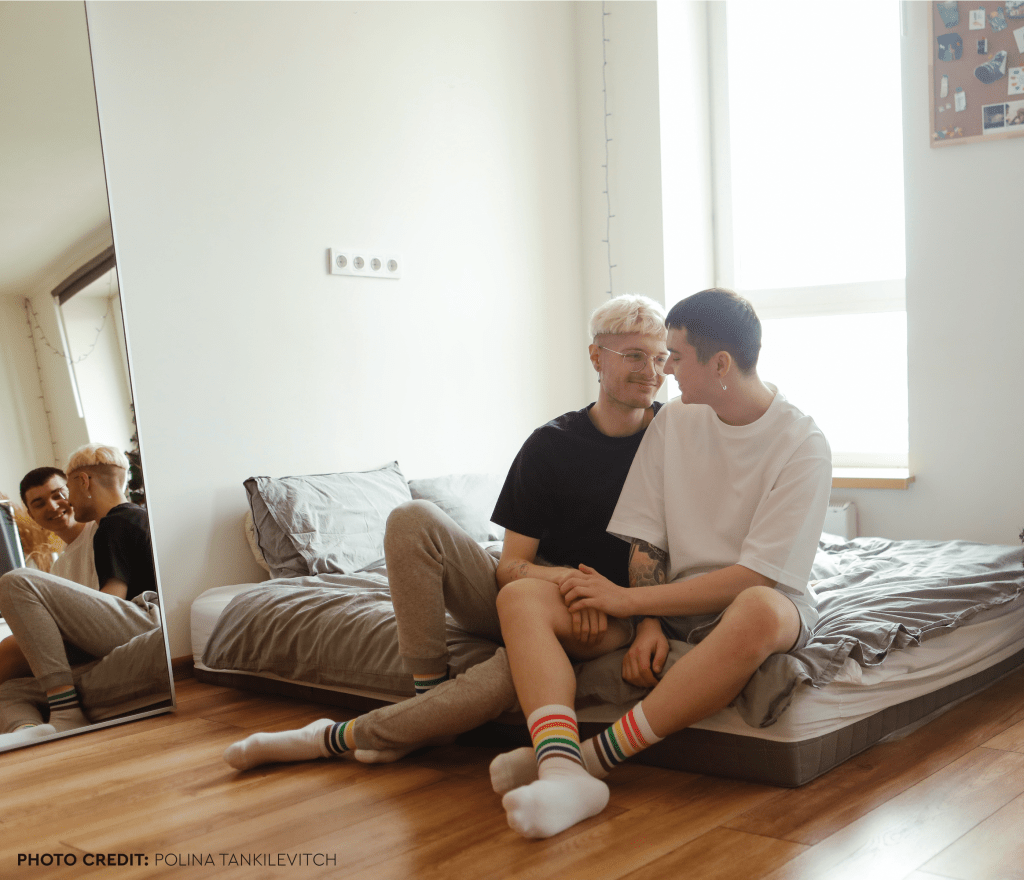 Two men sitting on a mattress both wearing rainbow socks. Photo credit Polina Tankilevitch
